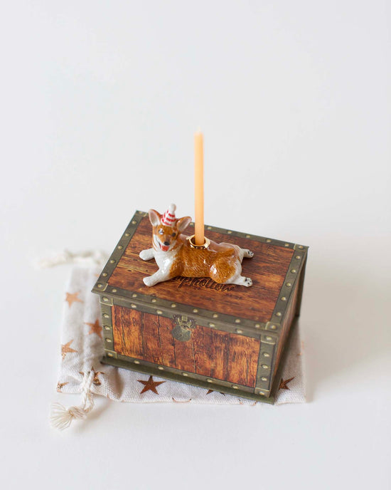 A hand-painted wooden music box with a ceramic figurine of a cat and a candle on top, placed on a white surface with a patterned cloth underneath, featuring the Camp Hollow year of the dog cake topper.