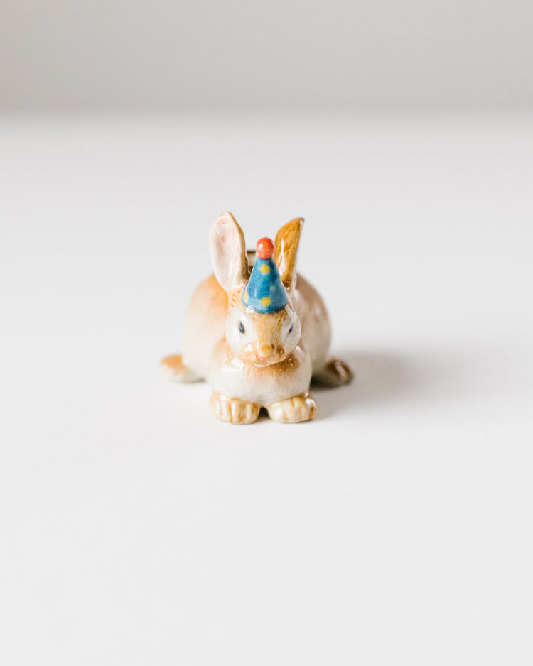 A small ceramic figurine of a rabbit, hand-painted in soft hues of brown and white, wearing a blue cap, displayed against a plain white background. This is the Camp Hollow Year of the Rabbit Cake Topper.