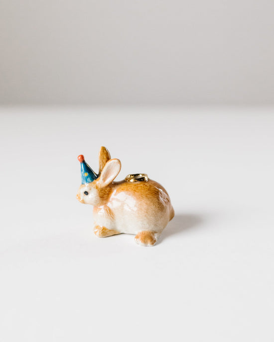 A small hand-painted porcelain Year of the Rabbit cake topper with a blue party hat, set against a plain white background by Camp Hollow.