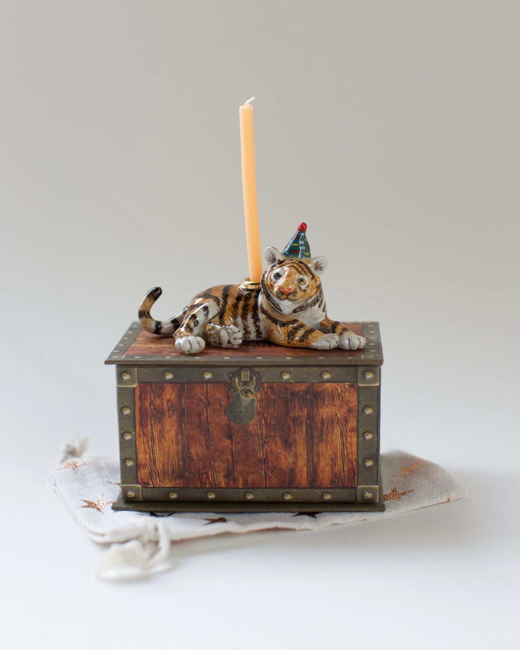 A Camp Hollow year of the tiger cake topper, with a party hat, lying atop an antique wooden chest with a single tall candle.