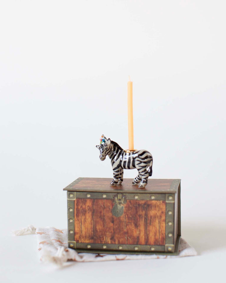 A hand-painted camp hollow zebra cake topper with a candle on its back stands on a small wooden chest against a plain, light background.