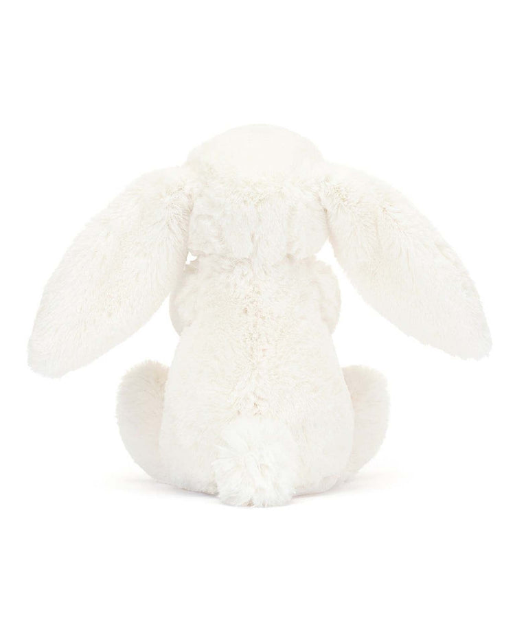 The bashful carrot bunny little by Jellycat is a seated plush toy bunny seen from behind, featuring long floppy ears hanging down its sides and a fluffy tail, all in white. This stuffed animal bunny clutches a tiny carrot.