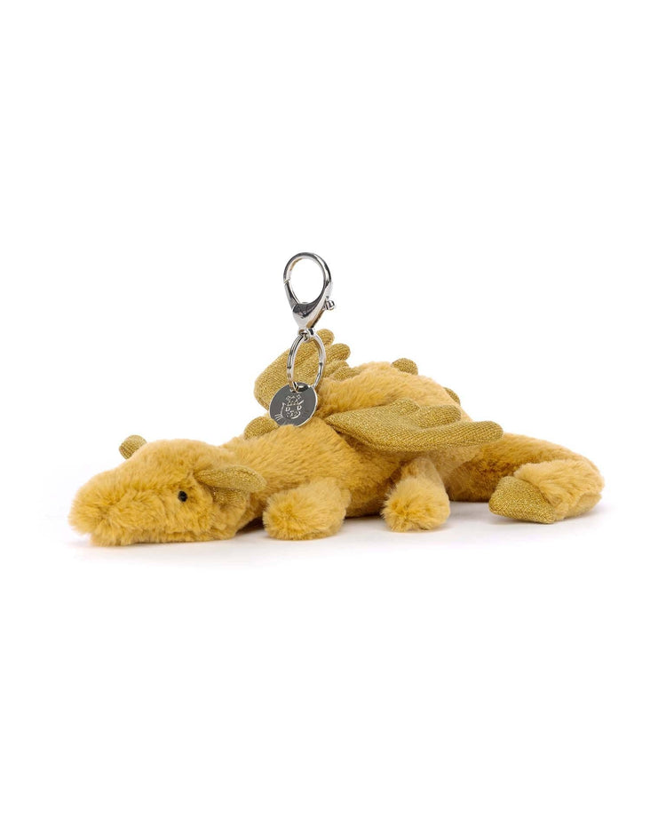 A golden dragon bag charm from Jellycat, featuring glittery wings, a metallic clasp, and a metallic tag, lies on a white surface.