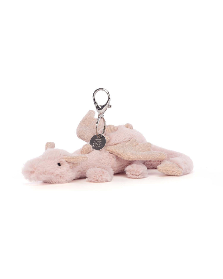 The Jellycat rose dragon bag charm, featuring glittery wings, a metallic clasp, and a small attached tag, sits against a white background. Perfect as a whimsical accessory for your bag!