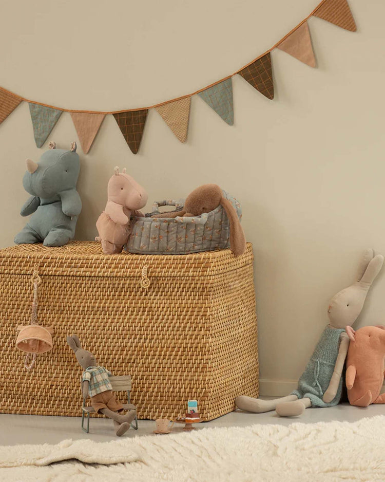 A maileg large carry cot in blue sits against a beige wall with a fabric pennant banner. Plush rabbits, animals, toy accessories, and a mini tea set complete the cozy nursery scene around the carry cot.