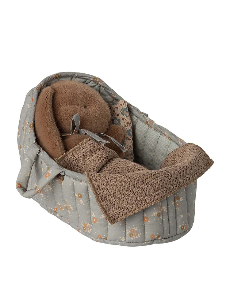A large blue maileg carry cot holds a brown stuffed bear, cushioned inside and covered with a crocheted brown blanket.