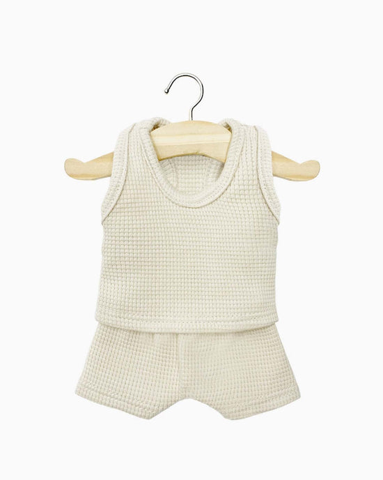 The Les P’tits Basiques waffle tank and short set in linen by Minikane, featuring a cream-colored, sleeveless woven top and matching shorts, is displayed on a wooden hanger against a white background. This set is perfect for Paola Reina Minikane dolls and inspires hours of imaginative play.