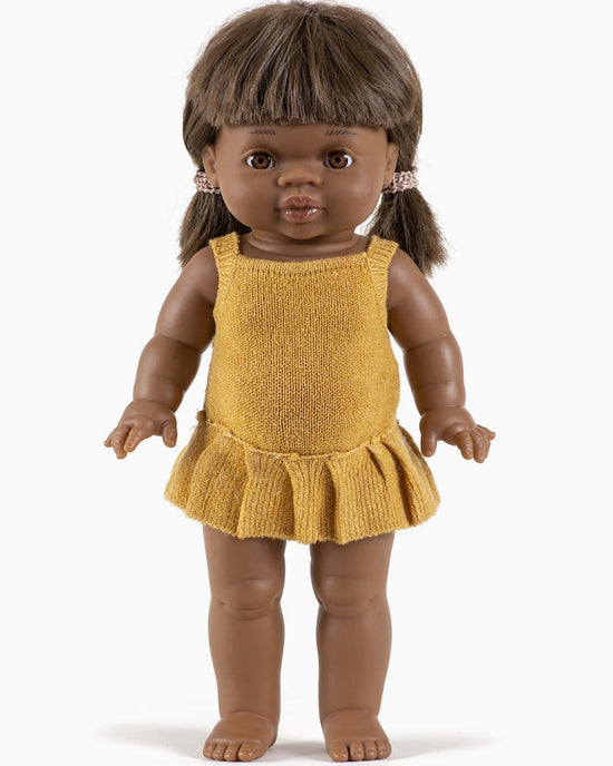 A brown-skinned doll with black hair in pigtails, dressed in an orléane knit romper in honey, stands upright with its arms slightly raised and eyes looking forward. This timeless doll captures the charm of Minikane dolls.