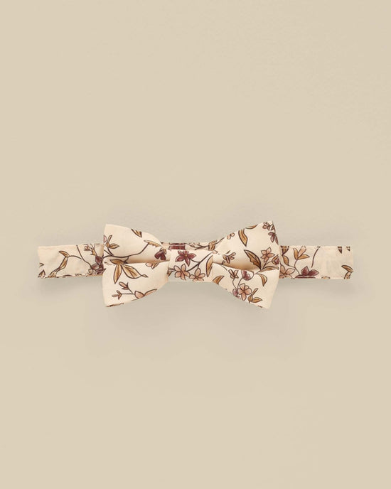 The "bow tie in fig floral" by noralee is crafted from 100% cotton, featuring a charming cream base embellished with a floral pattern of small red and brown flowers, making it perfect for quick and easy wear.