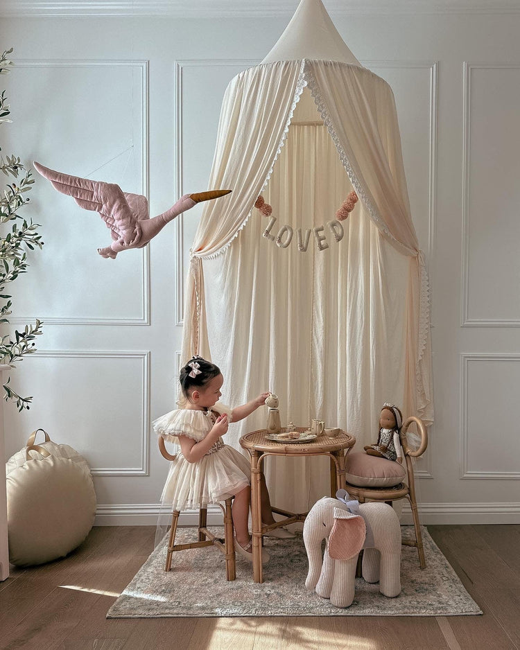 A young girl in a cream dress sits at the Poppie Kids Table by Poppie Toys under a canopy with a Loved banner, surrounded by stuffed animals in a softly lit, elegant playroom.
