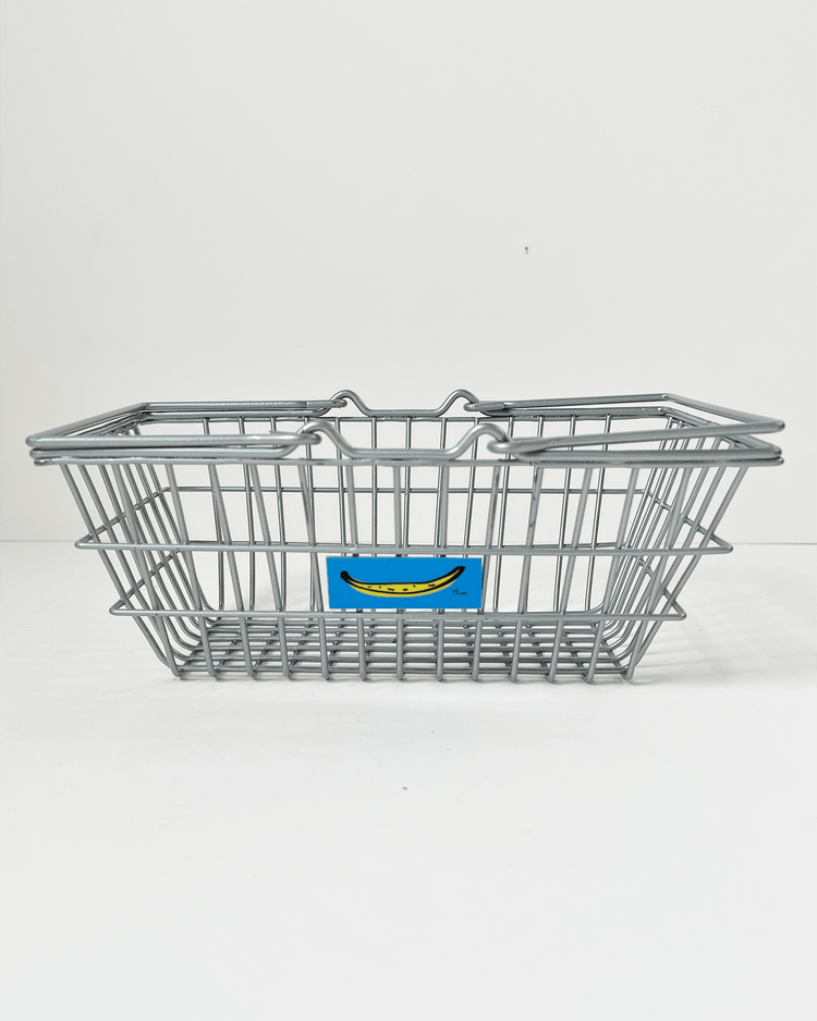 The teeny tiny market basket in silver, featuring a small blue banana sticker, is set against a plain white background. This charming play food item from teeny tiny market enhances any collection and promises hours of imaginative fun for children.