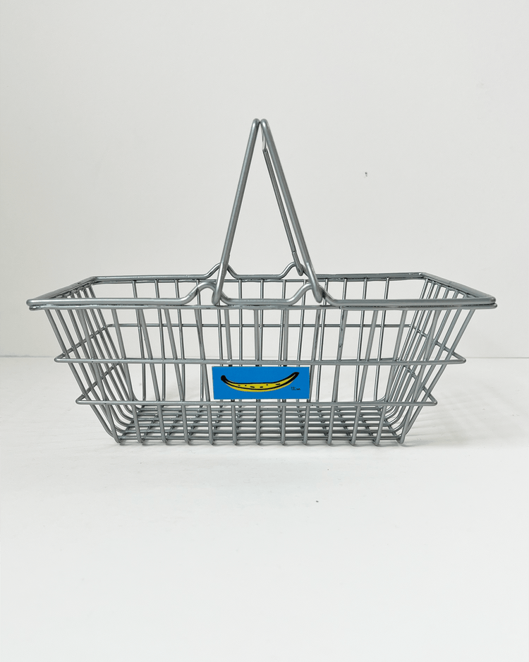 The teeny tiny market basket in silver, ideal for organizing child-safe toys, comes with a blue label showcasing a yellow smiley face, set against a white background.