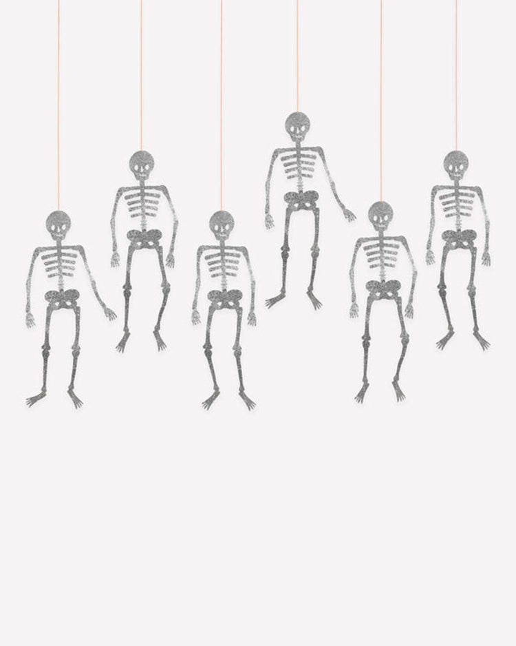 Six meri meri glitter jointed skeleton decorations hang in a row from thin strings against a plain white background. Each poseable skeleton features sparkling silver glitter and has its arms and legs arranged differently, creating a playful, festive display.