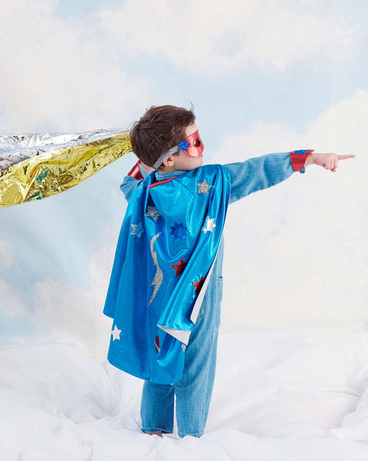 A child wearing the meri meri blue superhero costume—featuring a blue cape, red mask, and headband—stands on a bed with one arm extended forward, holding a gold and silver sheet behind them. The backdrop of a blue sky with clouds highlights the playful spirit and imaginative adventures inspired by this kids’ dress up set.