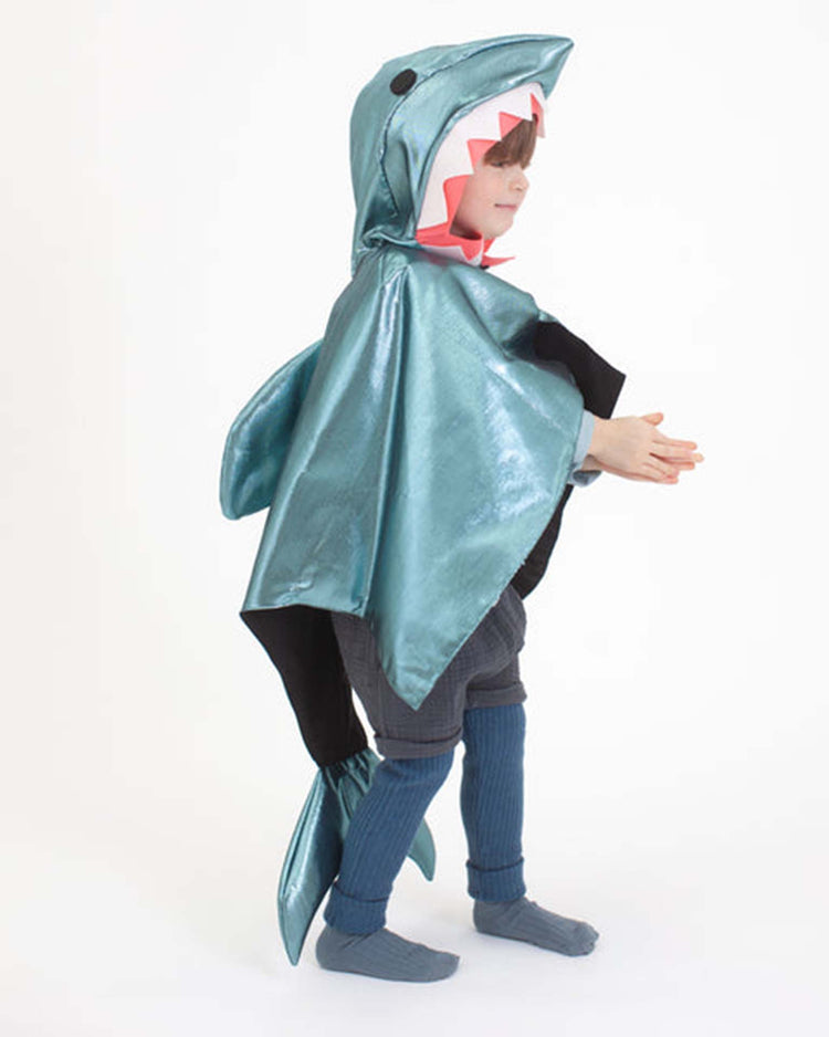 A child dressed in the meri meri shark costume, featuring a shiny blue finish with a shark fin and tail, stands sideways against a plain white background, hands together and looking forward.