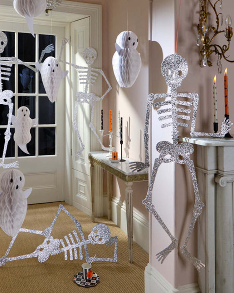 Silver foil skeleton and white ghost decorations hang in a room with a mirror, table, and fireplace. Orange and black candles on the mantel and table create a festive Halloween atmosphere that’s perfect for showcasing your favorite Halloween party decor, such as meri meri giant glitter jointed skeleton decorations.