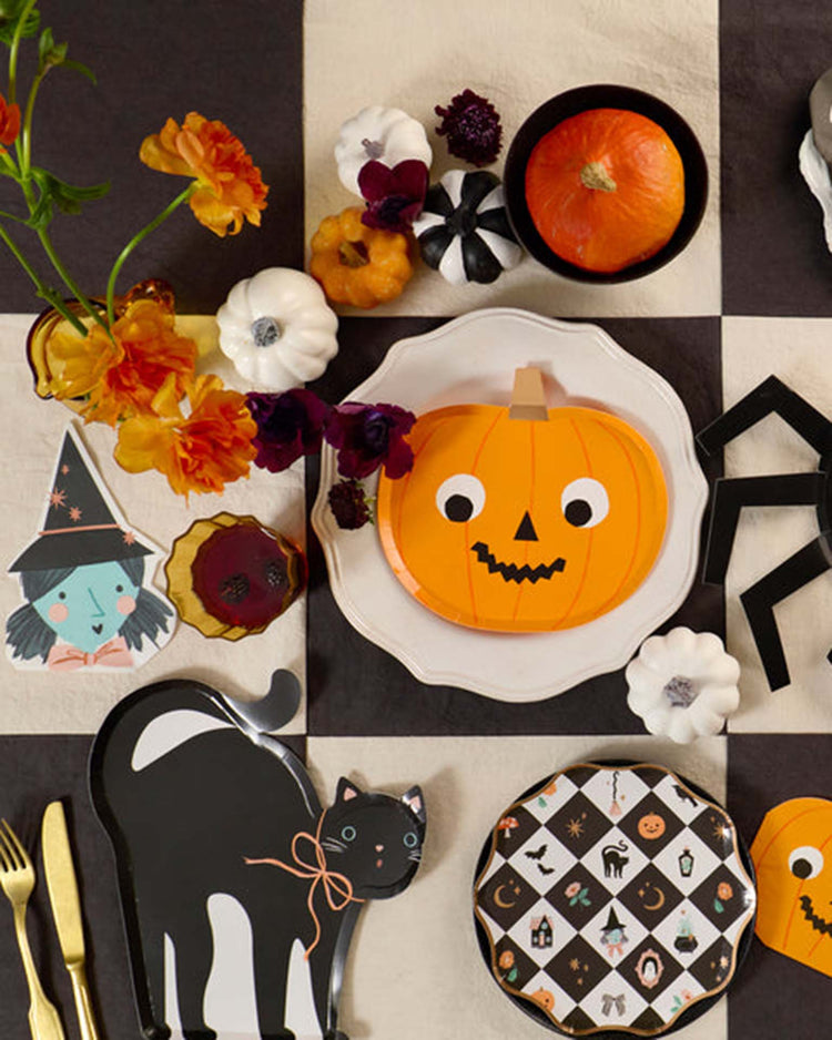 A Halloween-themed party table setting featuring pumpkin plates, a black cat plate, gold cutlery, flowers, small pumpkins, and meri meri witch shaped napkins—all arranged on a black-and-white checkered tablecloth.