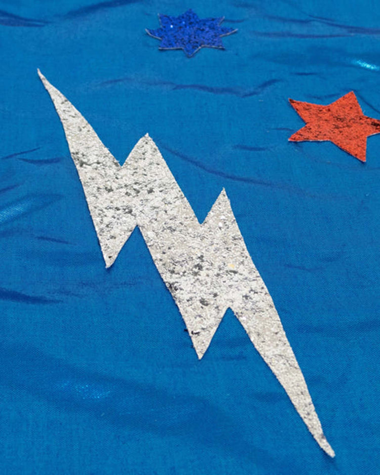 A sparkling silver glitter lightning bolt decorates the blue fabric of the meri meri blue superhero costume, with one blue and one red glitter star above it—making it perfect for inspiring kids' dress-up adventures as part of a superhero cape or Blue Superhero Dress Up Kit.