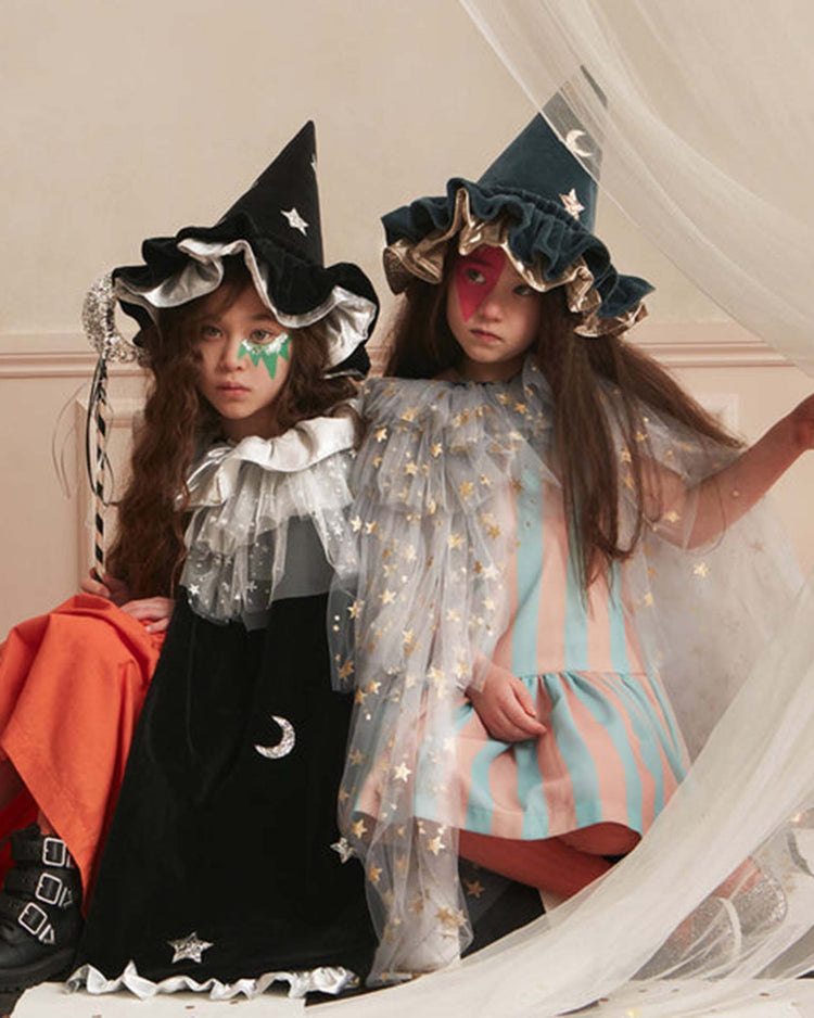 Two children in whimsical wizard costumes sit together; one wears the meri meri pointed blue hat and a black velvet cape adorned with silver stars, while the other sports a striped dress and hat sprinkled with gold stars—a perfect Halloween accessory. Both have face paint under their eyes. Sheer fabric drapes beside them.