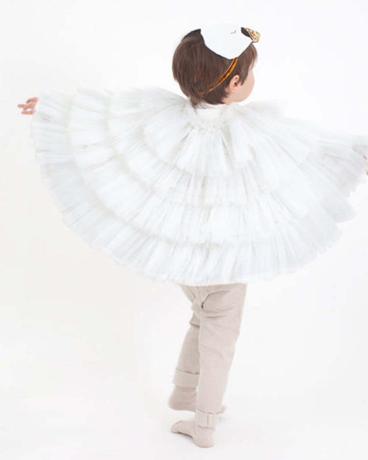 A young child stands with arms outstretched, facing away from the camera against a plain light background, wearing the meri meri swan cape dress up set—complete with a white mask and delicate tulle wings to capture the elegance of a swan.