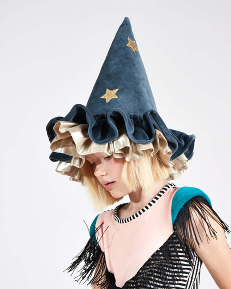 A child wearing the meri meri pointed blue hat—featuring ruffles, gold stars, and a colorful fringed top evocative of whimsical Halloween accessories—looks downward against a plain background.