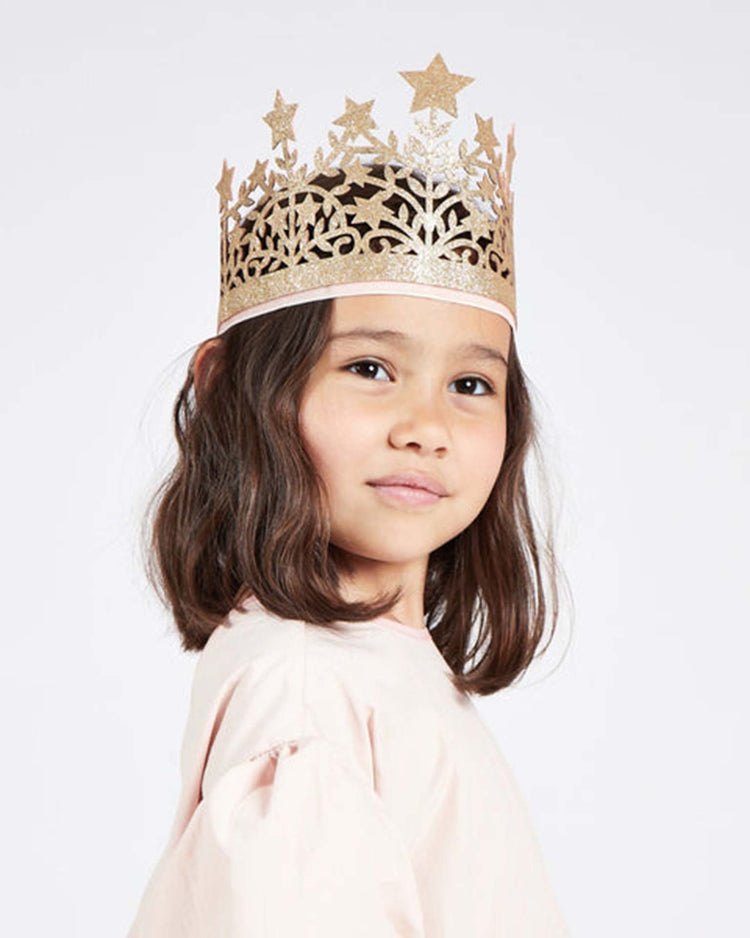 A young girl with shoulder-length brown hair, wearing a light pink top and the meri meri star crown—a decorative gold dress-up accessory adorned with stars—stands against a plain white background, looking confidently at the camera.