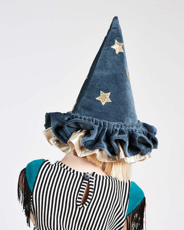 A child, seen from behind, wears the meri meri pointed blue hat—a whimsical accessory adorned with gold stars and a ruffled brim—paired with a black-and-white striped shirt featuring teal sleeves.