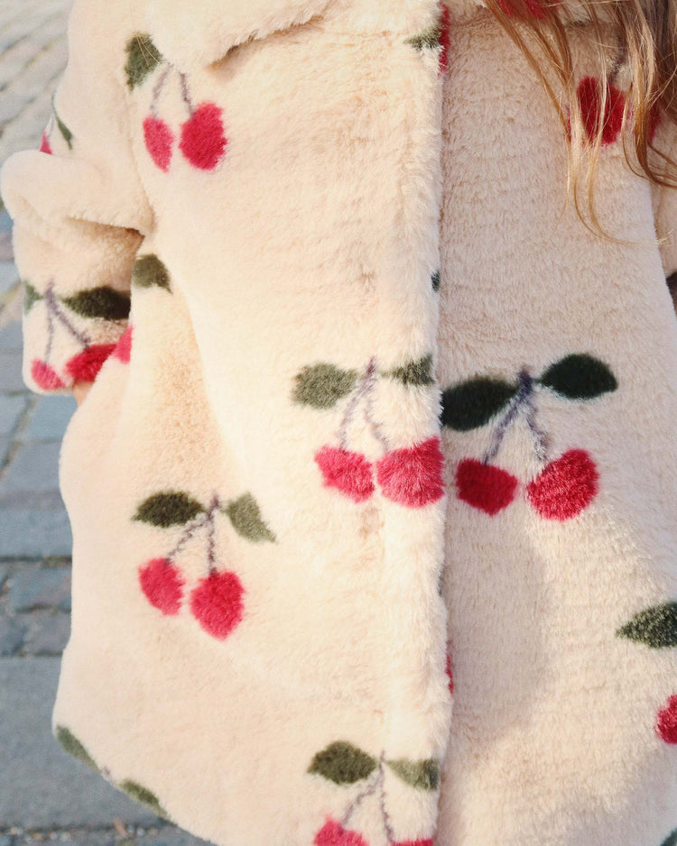A close-up of the konges sløjd fuzzie coat in ma grande cerise, a cream-colored plush coat adorned with a cherry pattern featuring red cherries and green leaves. The rounded collar is partially visible, along with part of a person’s arm and long hair. Crafted from GRS-certified polyester, the coat stands out against the stone pavement in the background.