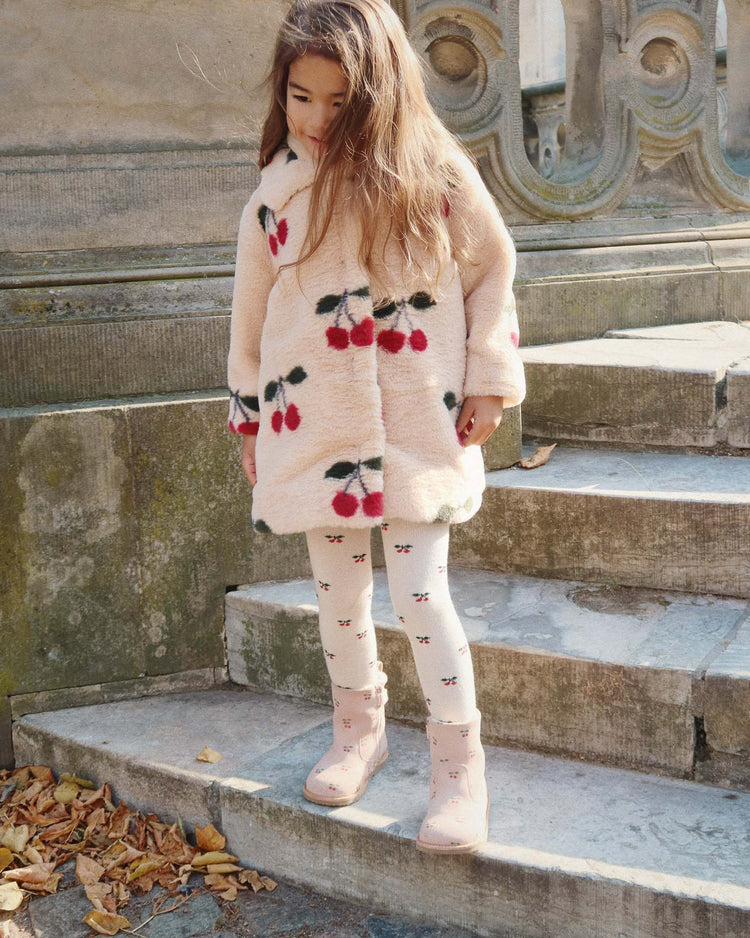 A young girl wearing the konges sløjd fuzzie coat in ma grande cerise, featuring a plush cherry pattern and rounded collar, with matching tights, stands on outdoor stone steps surrounded by fallen autumn leaves.