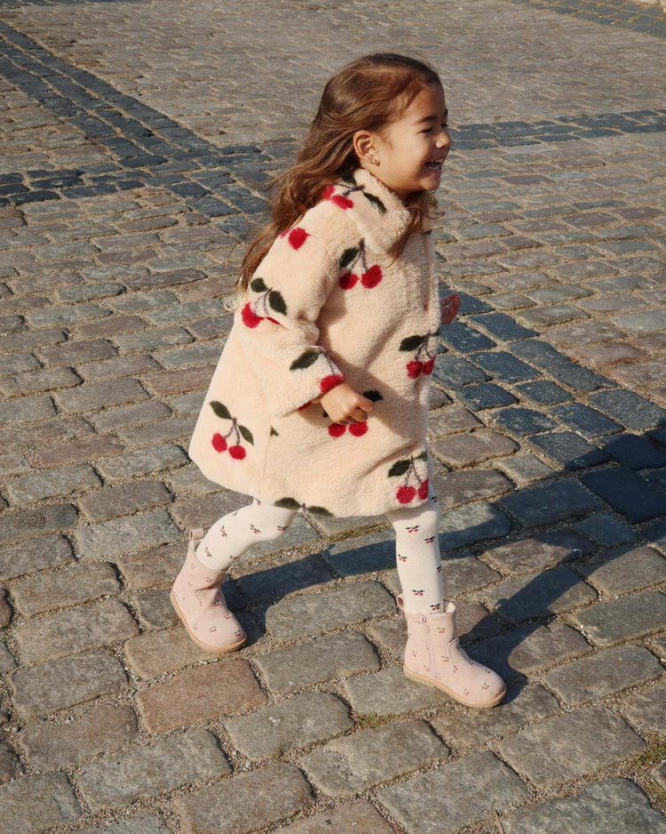 A young girl joyfully walks on a sunlit cobblestone path, wearing the konges sløjd fuzzie coat in ma grande cerise—a cream plush coat adorned with red cherry patterns and a rounded collar—paired with white tights and pink boots.
