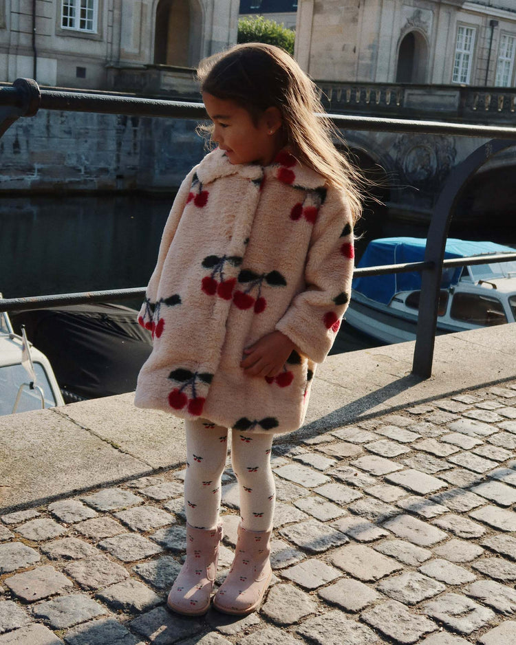 A young girl wearing the konges sløjd fuzzie coat in ma grande cerise, featuring cherry patterns and a rounded collar, stands on a cobblestone walkway by a canal with boats, looking to her left in sunlight.