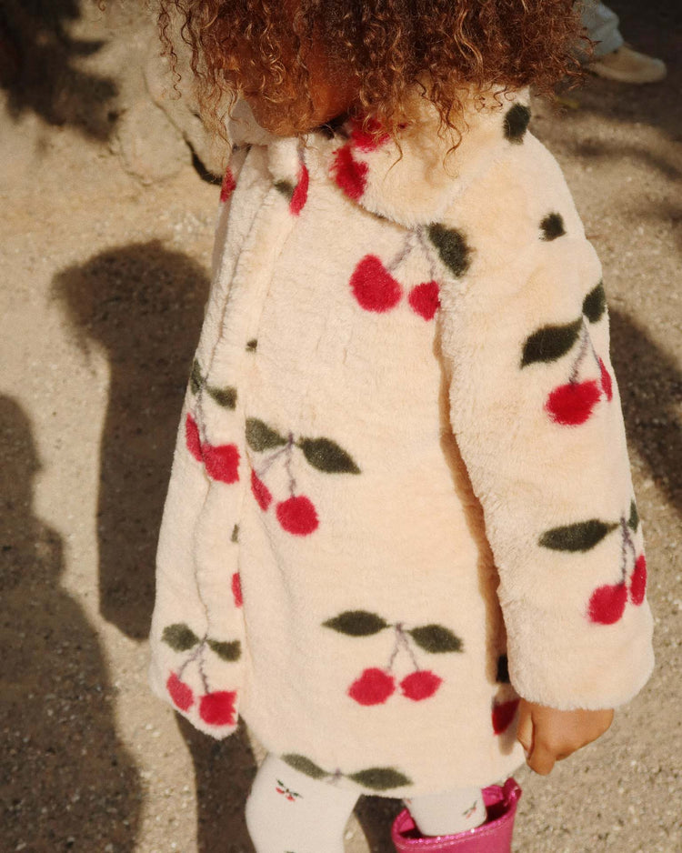 A young child with curly hair wears the konges sløjd fuzzie coat in ma grande cerise—a cream-colored plush coat crafted from GRS-certified polyester, adorned with red cherries and green leaves, and featuring a rounded collar—while standing outdoors on a sunny day.