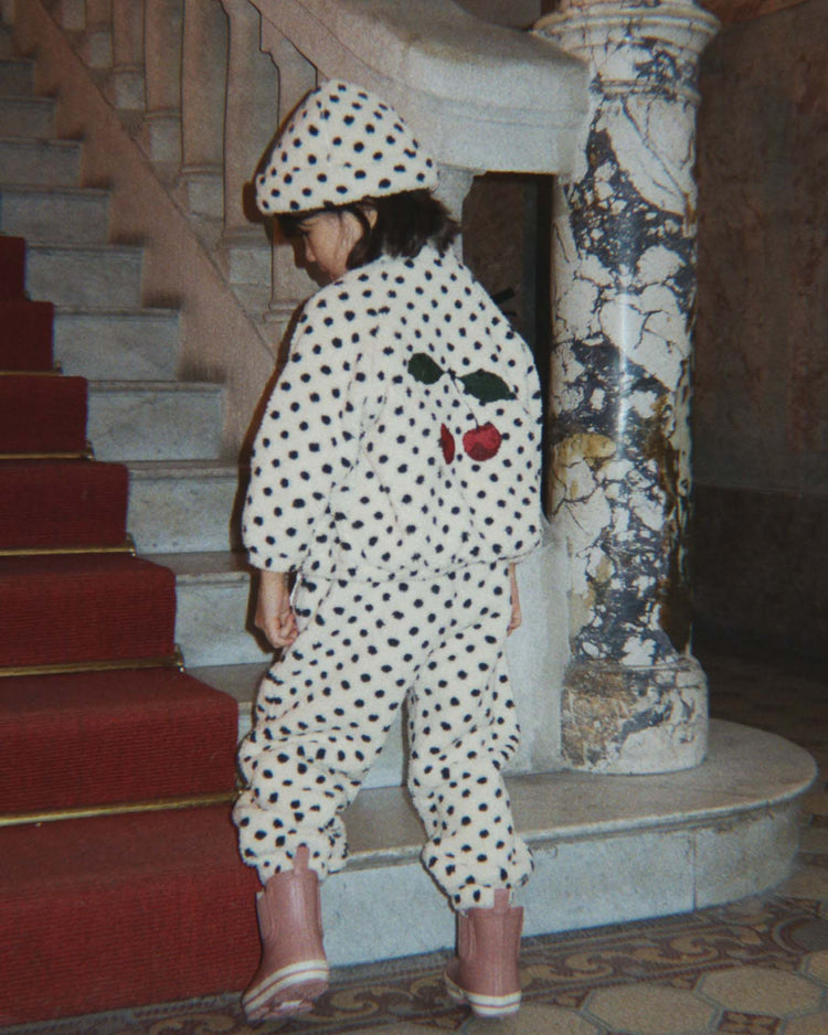 A child wearing the konges sløjd jody embroidery teddy jacket in dot lumiere—featuring a white, polka-dotted design and cozy recycled polyester fleece—stands on tiled flooring near red-carpeted stairs and a marble column.