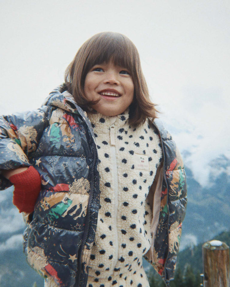 A smiling child stands outdoors against a backdrop of mountains and a cloudy sky, wearing the jody embroidery teddy jacket in dot lumiere by konges sløjd.