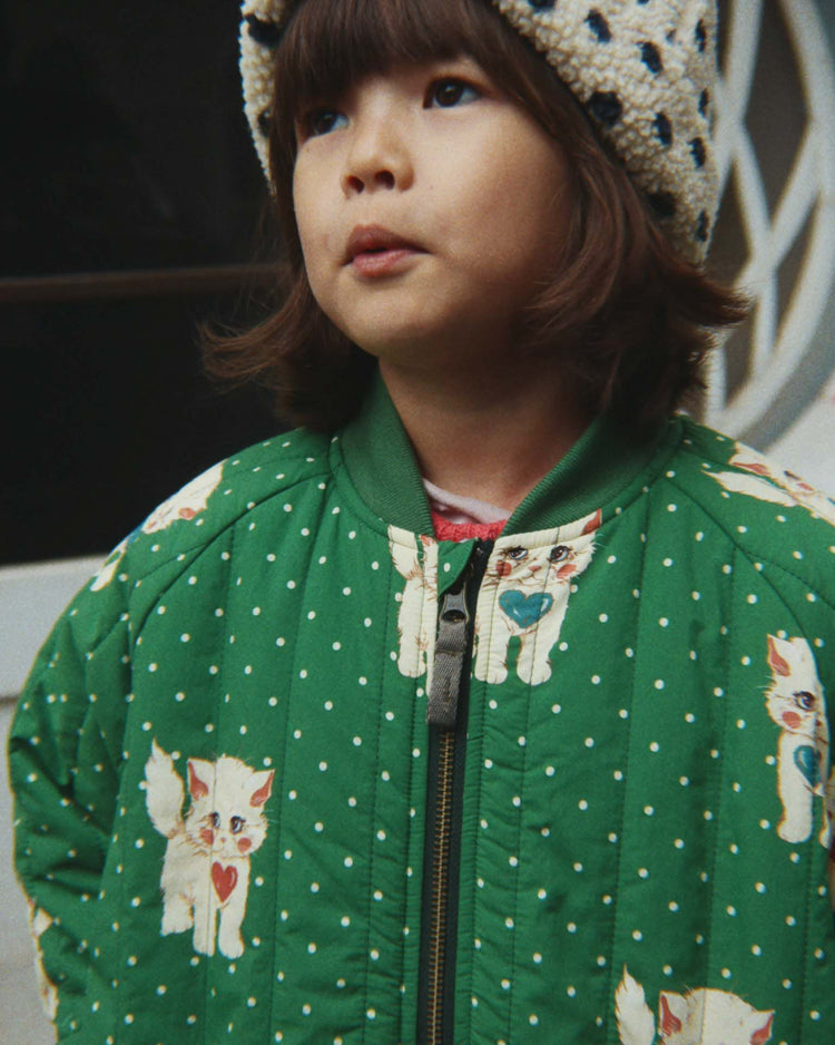 A young child wearing the konges sløjd juno printed bomber jacket in dot miao verde and a fuzzy hat stands outdoors, looking slightly upward.