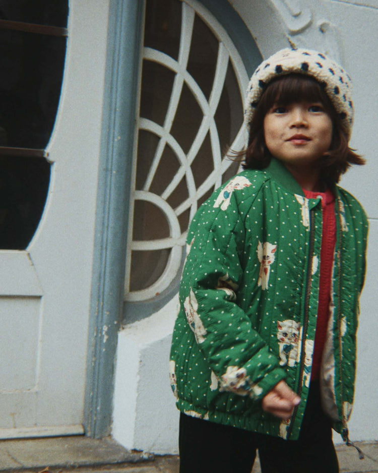 A young child wearing the konges sløjd juno printed bomber jacket in dot miao verde and a white hat stands outside near a decorative window and door, looking slightly toward the camera.