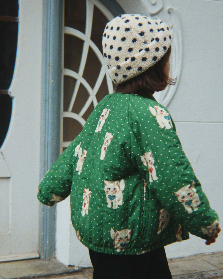 A child wearing the konges sløjd juno printed bomber jacket in dot miao verde and a white hat with black polka dots stands near a decorative window and white wall, facing away.