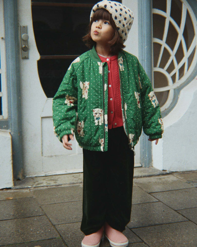 A young child wearing the konges sløjd juno printed bomber jacket in dot miao verde, along with a red sweater, dark pants, pink shoes, and a polka-dot hat, stands on a sidewalk looking up with a curious expression.