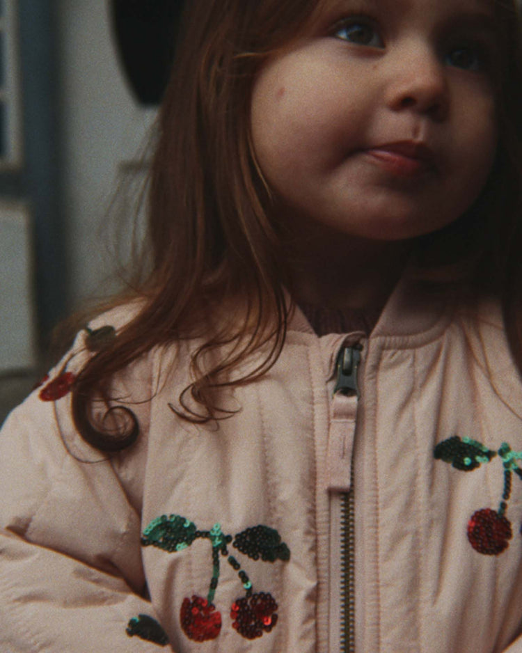 A young child with long brown hair wears the konges sløjd juno sequin bomber jacket in cameo rose, looking slightly upward with their tongue touching their upper lip.