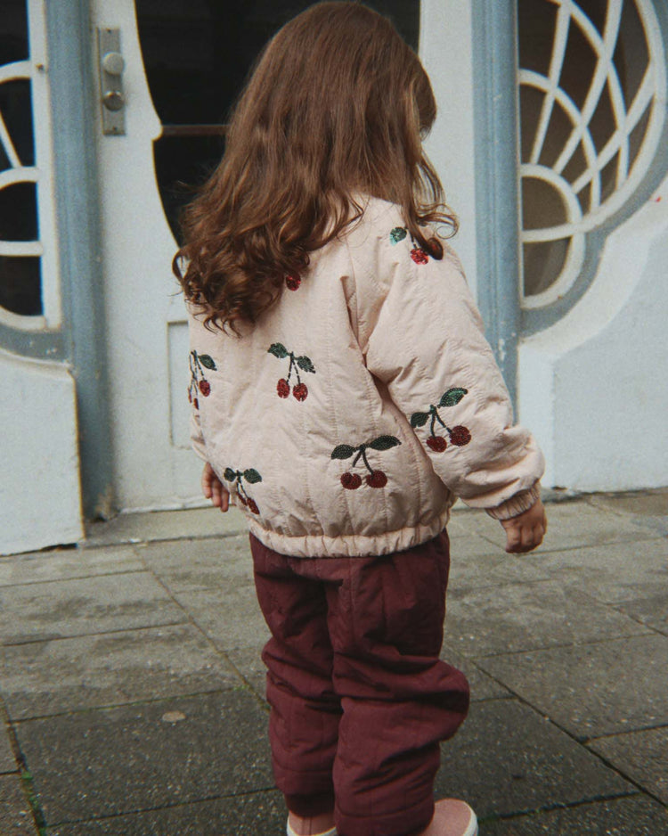A young child with long brown hair stands outside, facing away, wearing the Konges Sløjd Juno Sequin Bomber Jacket in Cameo Rose and maroon pants. This lightweight outerwear features water-repellent fabric, making it perfect for play on the gray sidewalk in front of a blue and white building adorned with decorative windows.