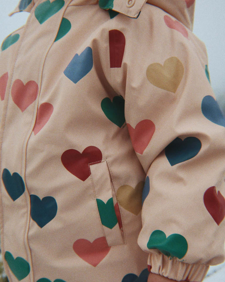 A close-up of a child wearing the Konges Sløjd "rain snowsuit in bon coeur coloré," a light pink, fleece-lined jacket adorned with large, colorful heart shapes in red, blue, yellow, and green. Only the torso and part of one arm are visible.