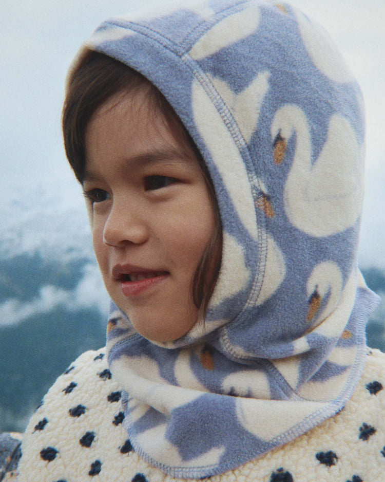 A young child wearing the konges sløjd tavi fleece balaclava in swan, paired with a cream sweater adorned with dark spots, stands outdoors against a backdrop of blurred mountains, perfectly capturing the charm of this cozy accessory.