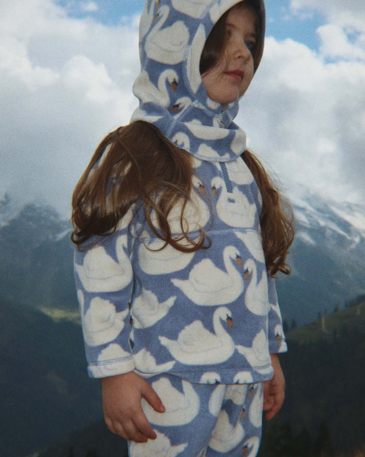 A young child wearing the Konges Sløjd tavi fleece balaclava in swan stands outdoors with mountains and a cloudy sky in the background.
