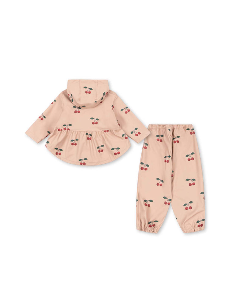 The konges sløjd faire rain set in ma grande cerise blush is a two-piece toddler outfit featuring a light pink hooded top and matching pants, both adorned with a red cherry print and elastic cuffs. Crafted from lightweight, water-repellent fabric to help keep little ones dry, the set is displayed laid flat on a white background.