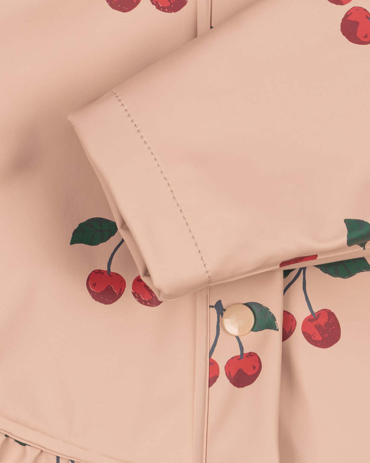 Close-up of the faire rain set in ma grande cerise blush by konges sløjd, showcasing a light pink, water-repellent fabric adorned with a cherry print featuring green leaves and red cherries. A folded sleeve with button detail appears in the foreground, emphasizing the lightweight, wind-resistant design of this stylish rain set.