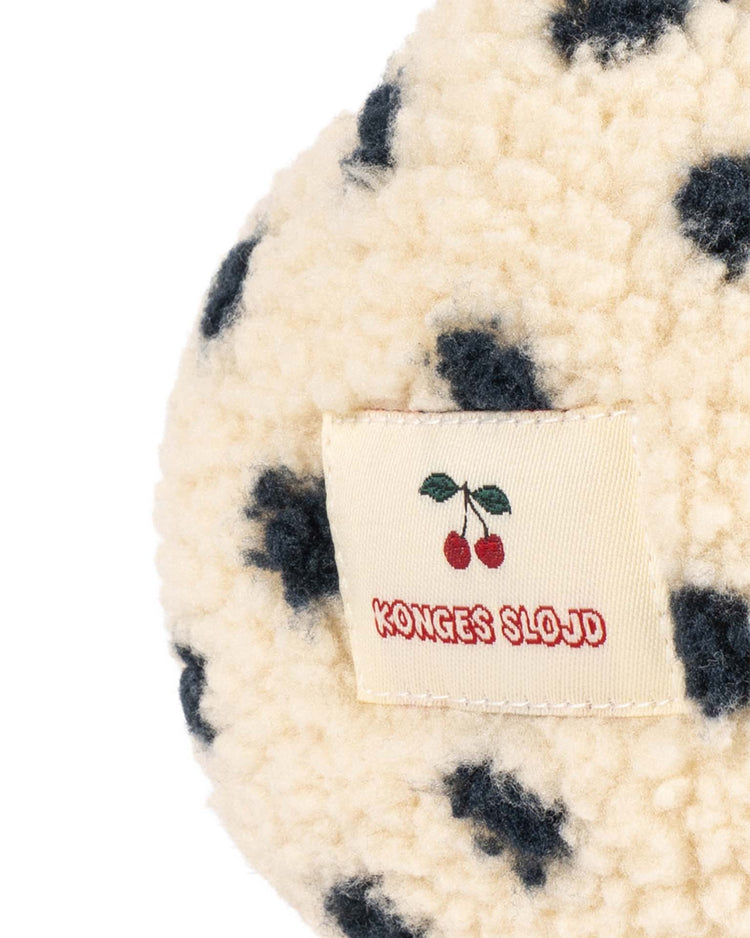 A close-up of the fluffy cream and black spotted fabric of the jody teddy earmuffs in dot lumiere by konges sløjd, OEKO-TEX certified for safety and comfort, featuring a sewn label with two cherries and "Konges Slojd" in red text.