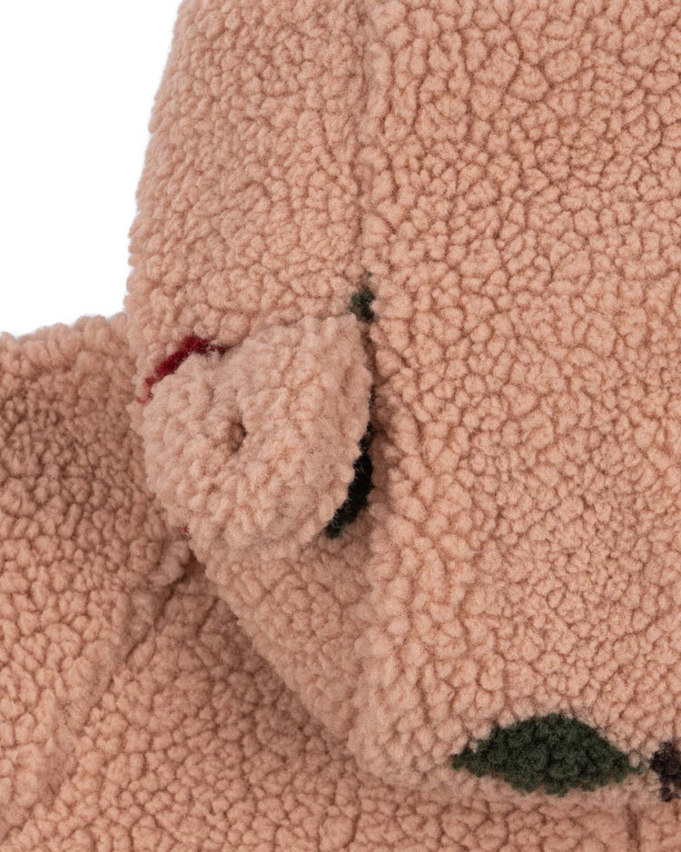 Close-up of the konges sløjd jody teddy onesie in ma grande cerise blush—a soft, tan-colored design crafted from textured, fleece-like fabric reminiscent of a cozy baby onesie. The image captures part of the rounded ear and head, accented by small dark green and red patches that highlight its recycled polyester construction for an eco-friendly touch.