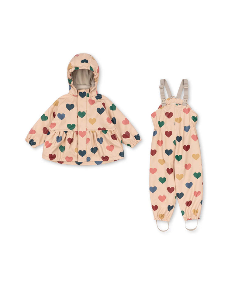 The rainy palme set girl in bon coeur coloré by konges sløjd features a children's waterproof raincoat and overalls in light beige adorned with a colorful heart pattern. The wind-resistant raincoat includes a hood, while the overalls offer adjustable straps, foot loops, and a soft fleece lining for added comfort.