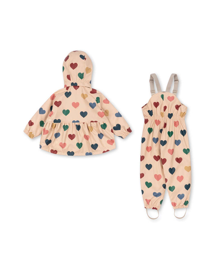 The "rainy palme set girl" in bon coeur coloré by konges sløjd features a beige waterproof and wind-resistant rain jacket with a hood, paired with matching overalls. Both pieces are adorned with a colorful heart pattern in red, blue, green, and pink, and are lined with cozy fleece for extra warmth on rainy days.