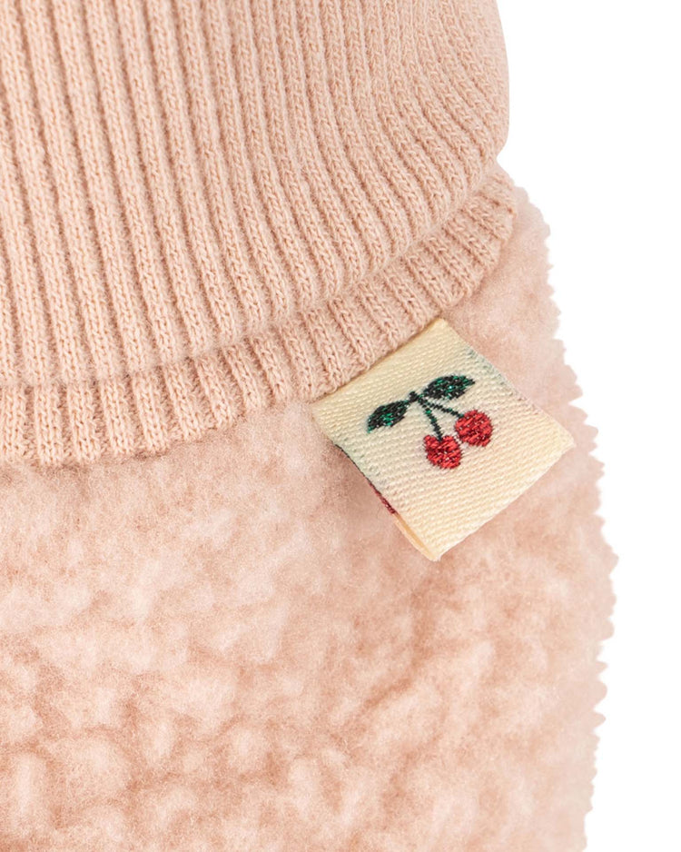 Close-up of the safa boots in rose smoke by konges sløjd, showcasing the soft pink textured fabric with a ribbed cuff and a small cream-colored tag adorned with an illustration of two red cherries and green leaves. The cozy material suggests a plush organic cotton lining, offering enhanced comfort and sustainable style.